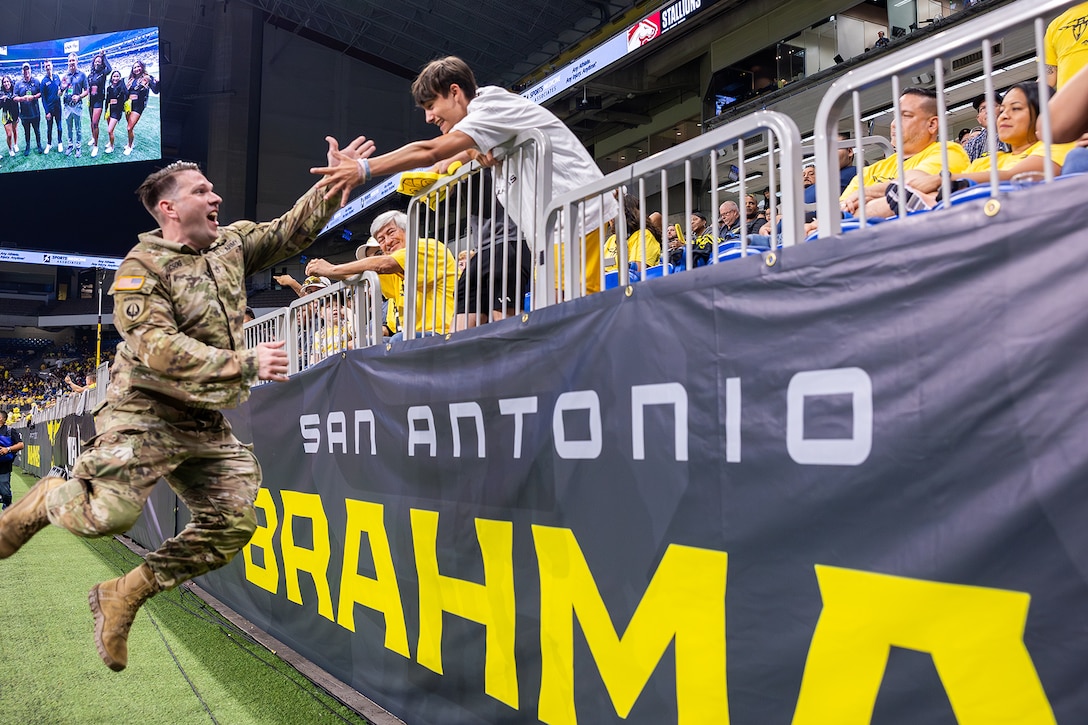 A soldier jumps from the field to greet a fan leaning down from the stands at a football game.