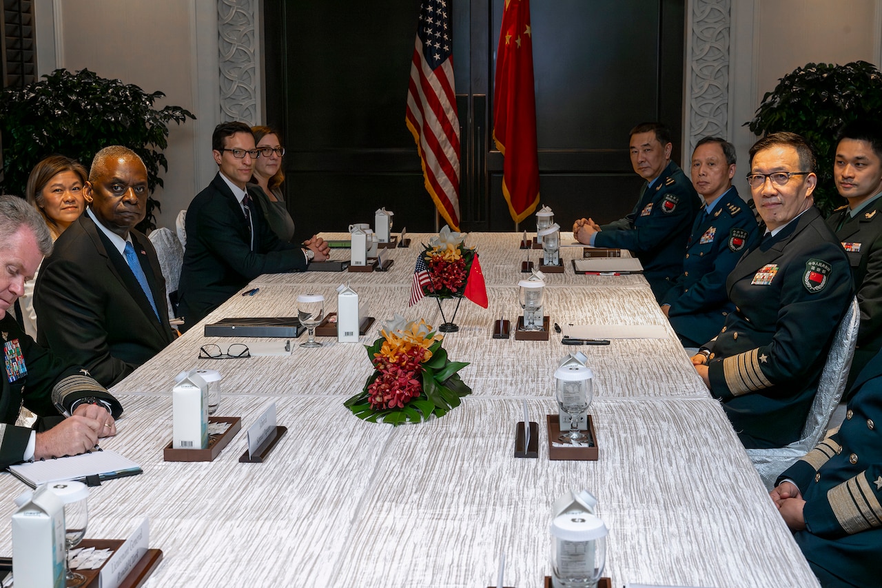 A group of people in business attire and military uniforms sit around a conference table.