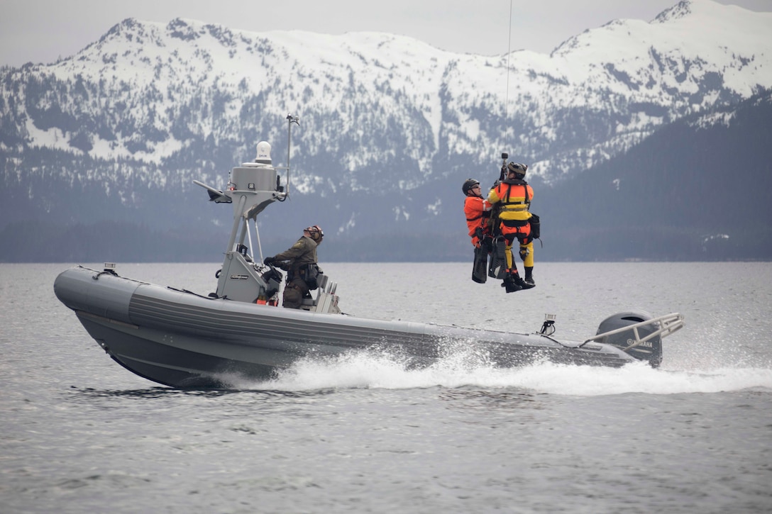 Two guardsmen in orange are hoisted from a small boat in a body of water with snowy mountains in the background.