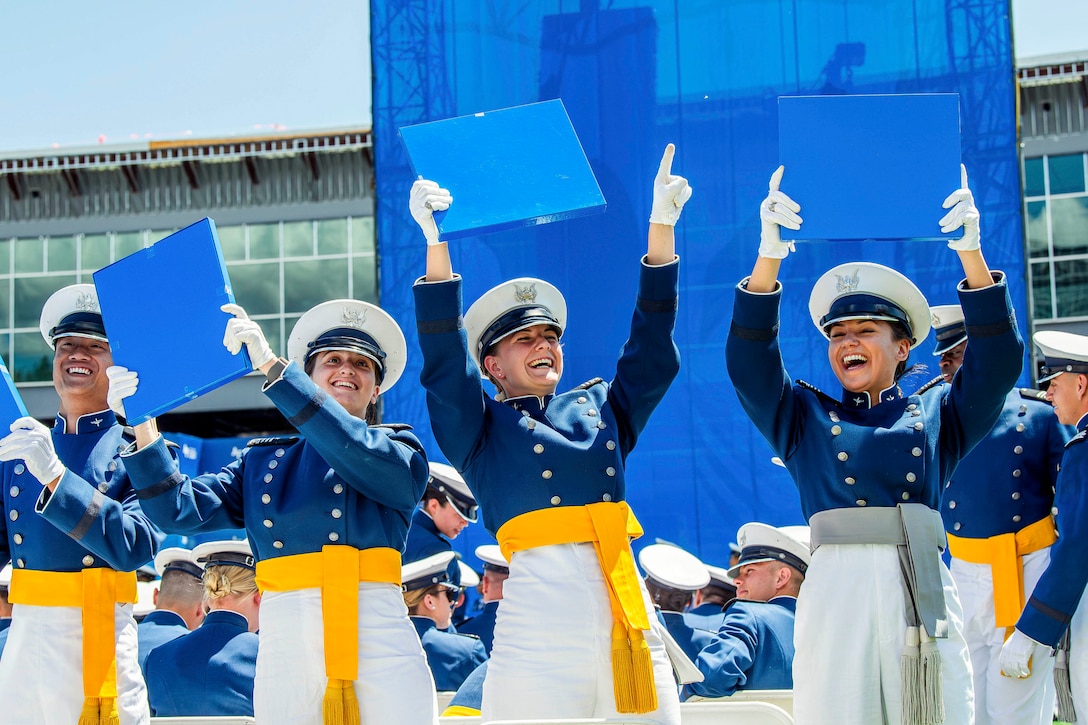 Four cadets in ceremonial dress smile while holding their diplomas up as dozens of fellow cadets sit behind them.