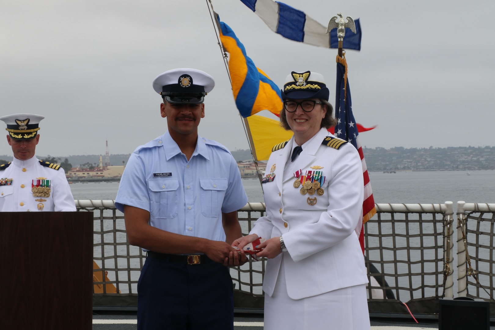 Seaman Fabio Caciel-Reyes (left), the most junior member of the U.S. Coast Guard Cutter Munro’s (WMSL 755) crew, presents Capt. Rula Deisher (right) with a commissioning pennant during Munro’s change of command ceremony in San Diego, May 30, 2024. Vice Adm. Andrew J. Tiongson, Commander, U.S. Coast Guard Pacific Area, presided over the ceremony in which Capt. James O’Mara relieved Deisher as Munro’s commanding officer. U.S. Coast Guard photo by Ensign Samika Lewis.