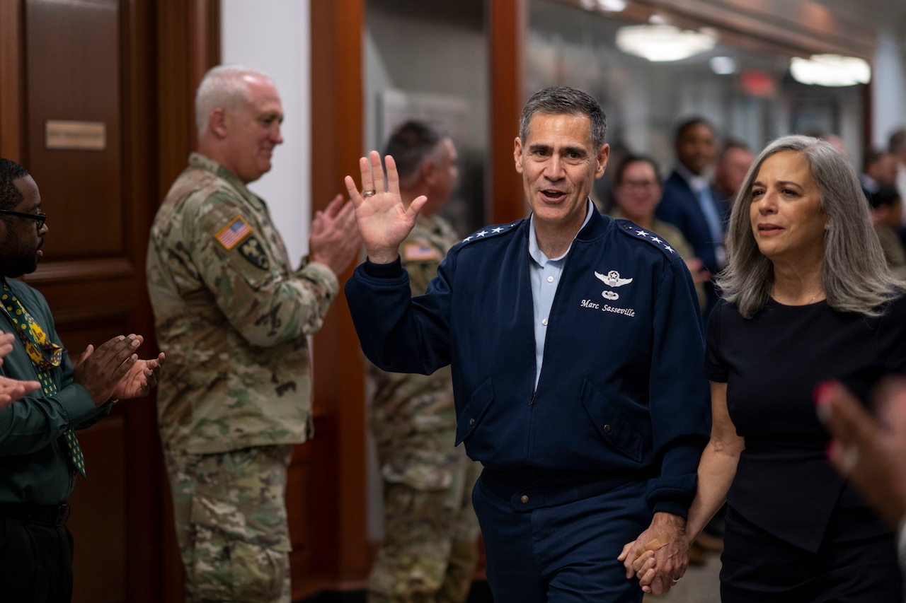 A man in military uniform waves and walks down a hall while holding the hand of a woman as people, some in military uniform, clap for them.