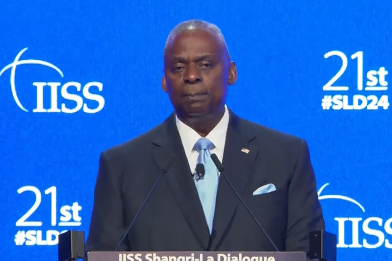 A person in a business suit speaks at a podium with a blue wall in the background that reads "IISS."