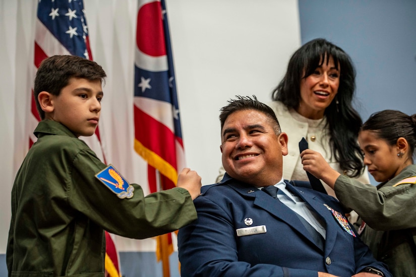 Two children pin new insignia on an officer sitting in a chair and smiling. A civilian stands behind them.