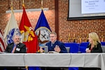 Three individuals sit behind a table on a stage as part of a panel.