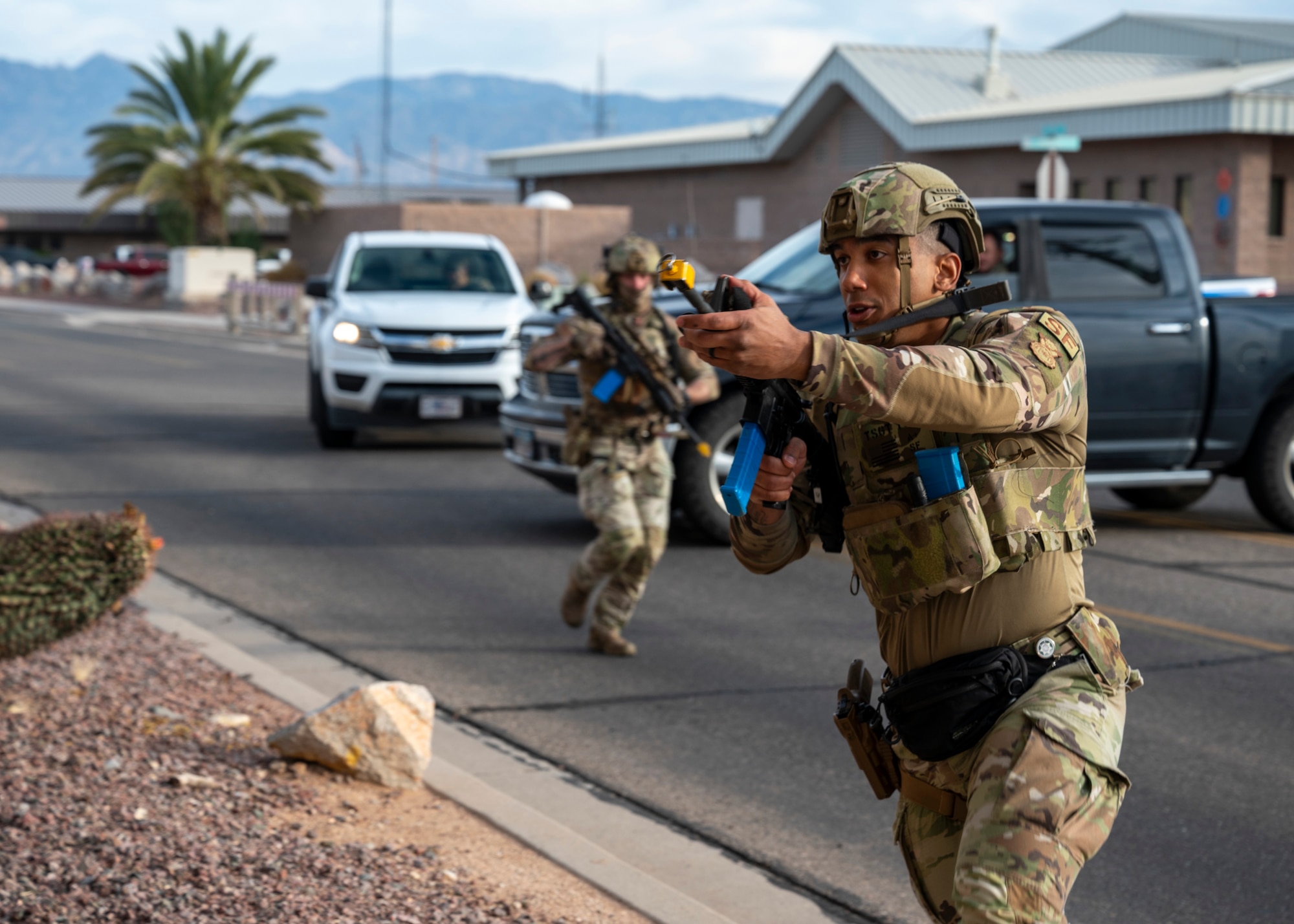 A man in military clothing holding a rifle runs, while another man in military clothing holding a rifle follows close behind.