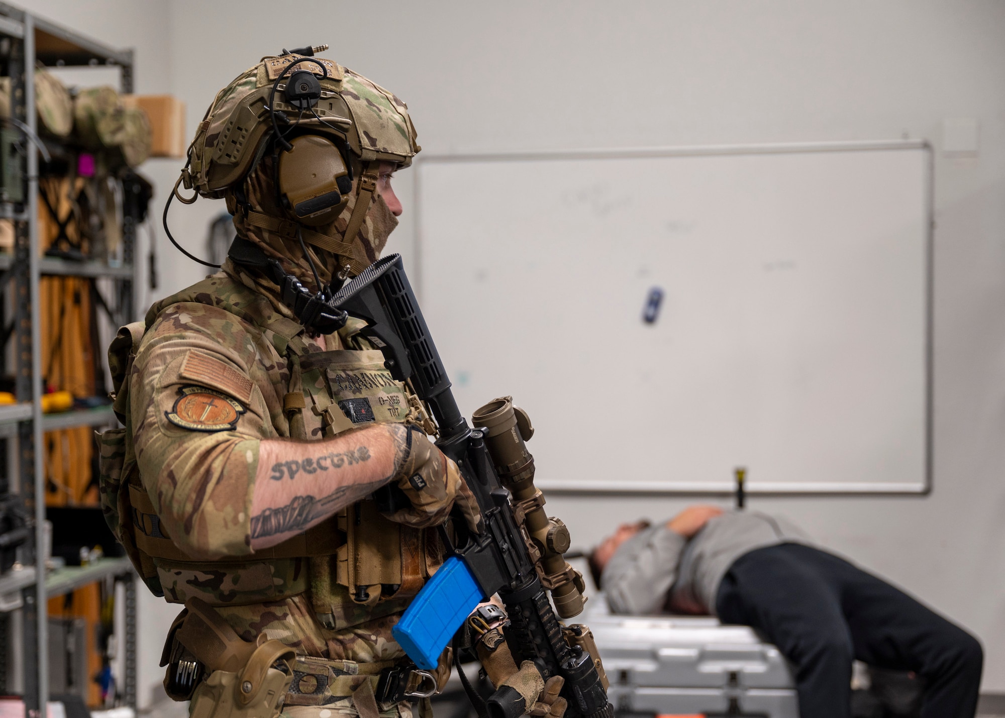A man in military clothing holds a rifle and looks across a room while a man in a sweatshirt and sweatpants is lying on the ground in the background.