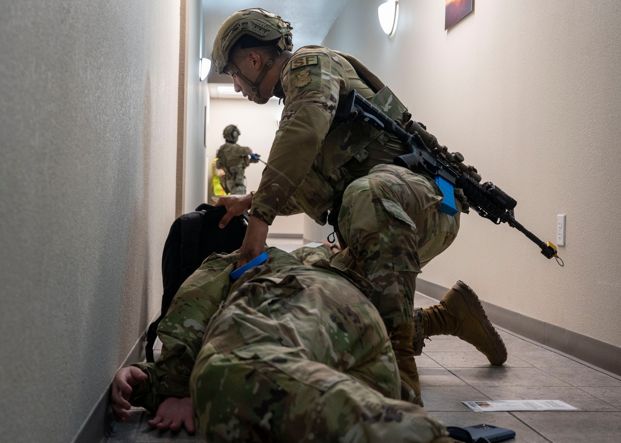A man in military clothing kneels overtop of another man in military clothing while another man in military clothing clears a hallway with a rifle.