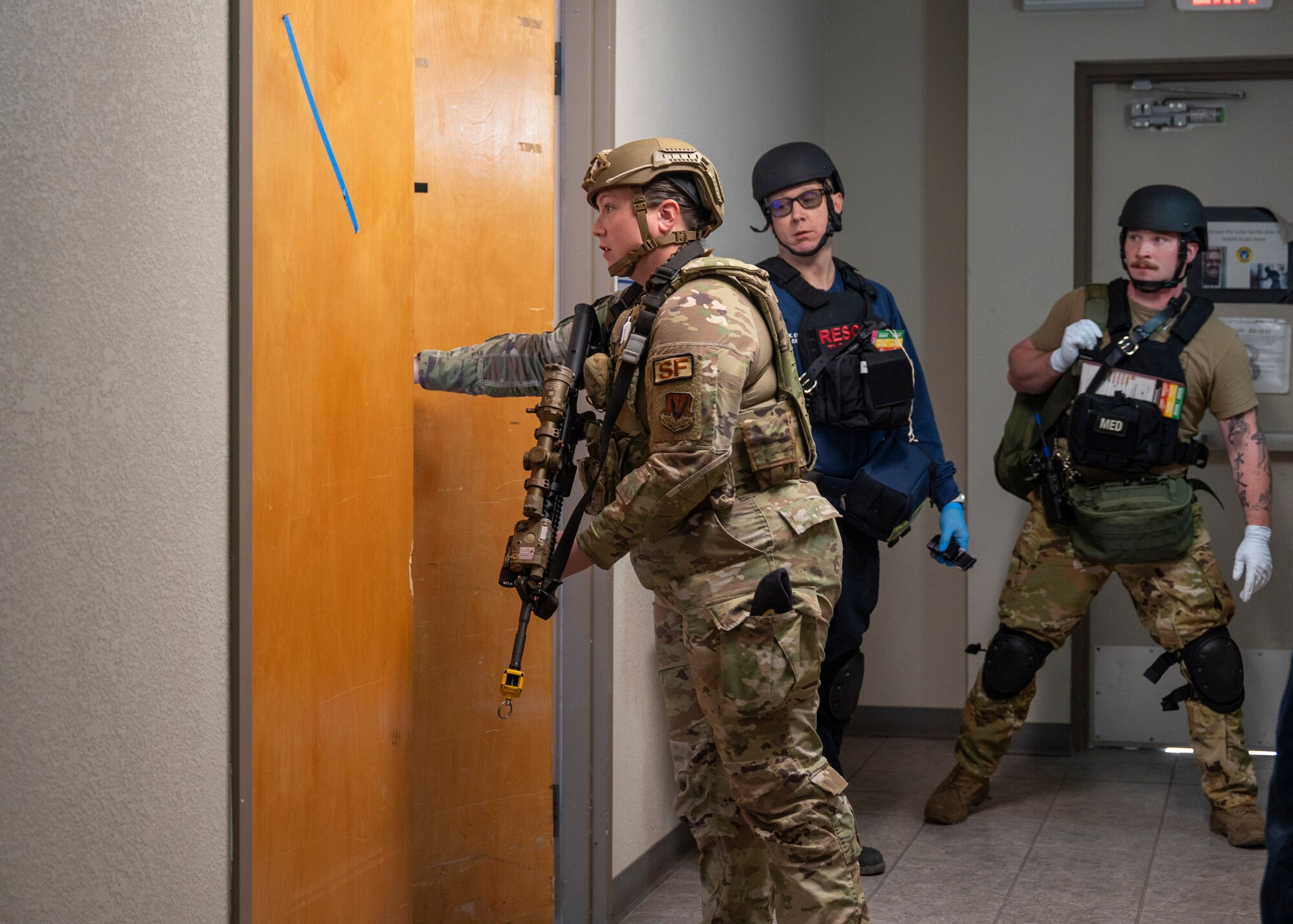 A woman in military clothing holding a rifle opens a door as two men in military clothing stand behind.