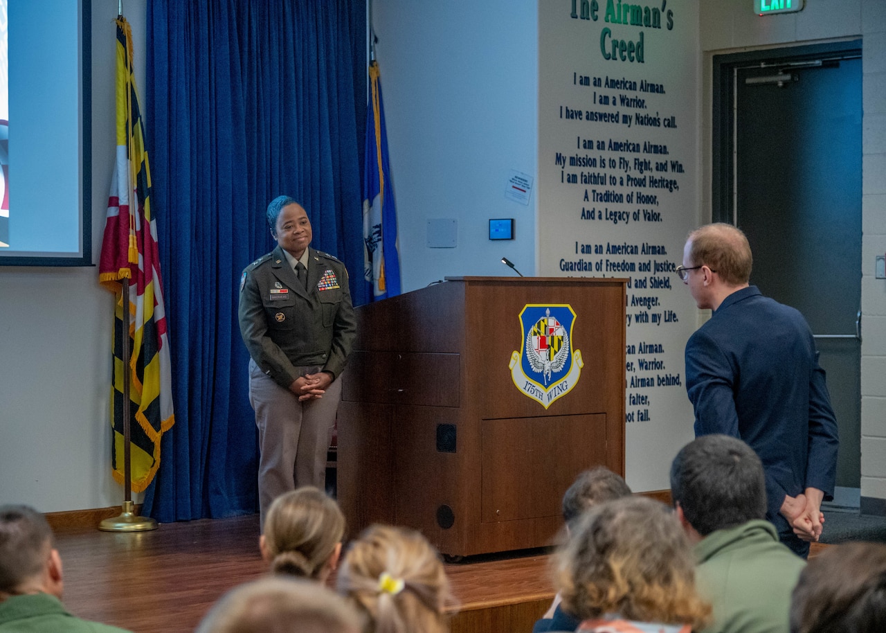 A woman in military uniform standing on a stage, smiles at a standing man in the audience.