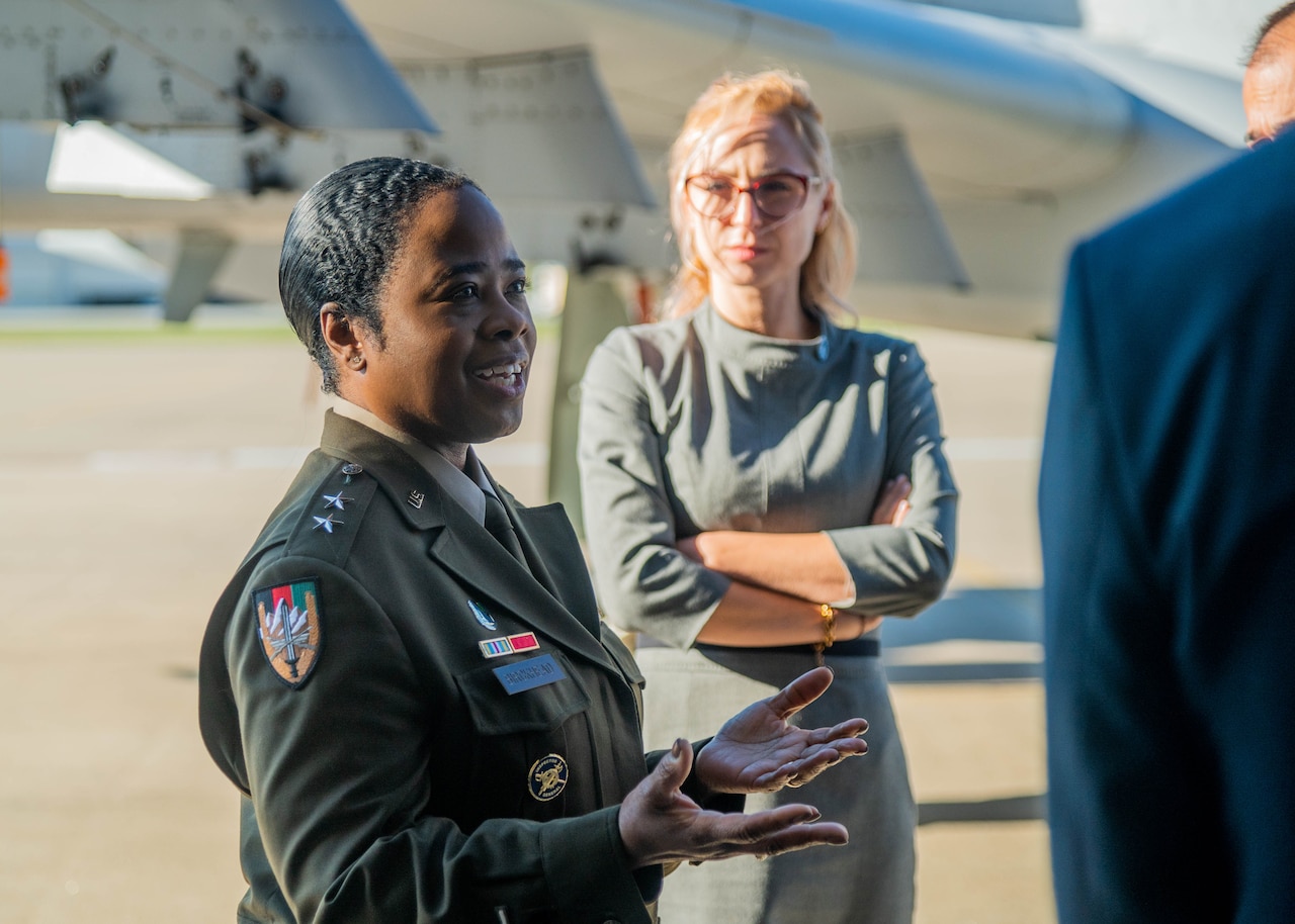 A woman in military uniform gestures with her hands open. Another woman in the background looks on.