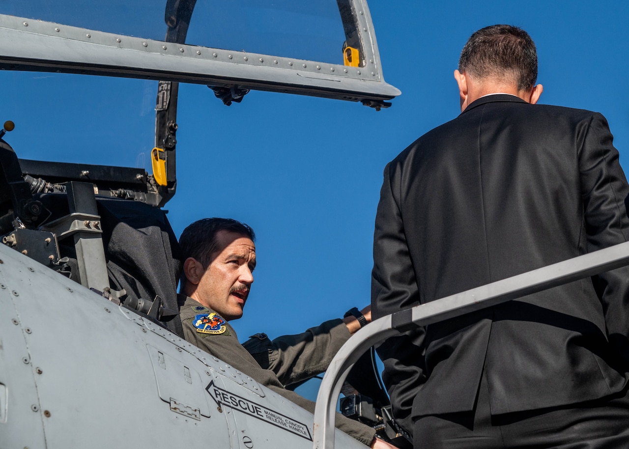 A military pilot sits in an aircraft speaking to a man in a business suit.