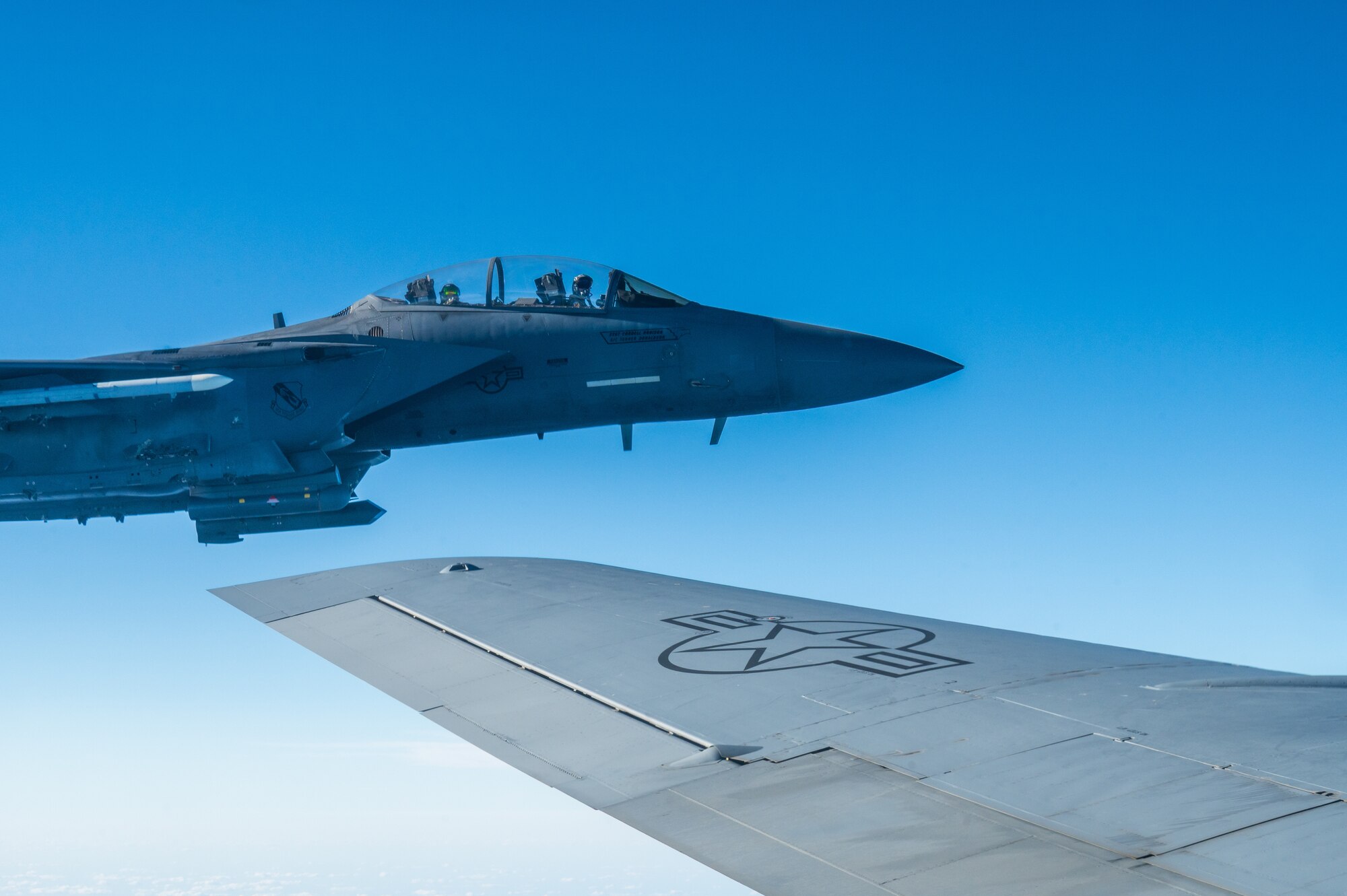 A military jet flies in formation with another military aircraft. The cockpit of one of the jets is visible at left, the wing of the aircraft from which the photo is taken is also visible (at right).