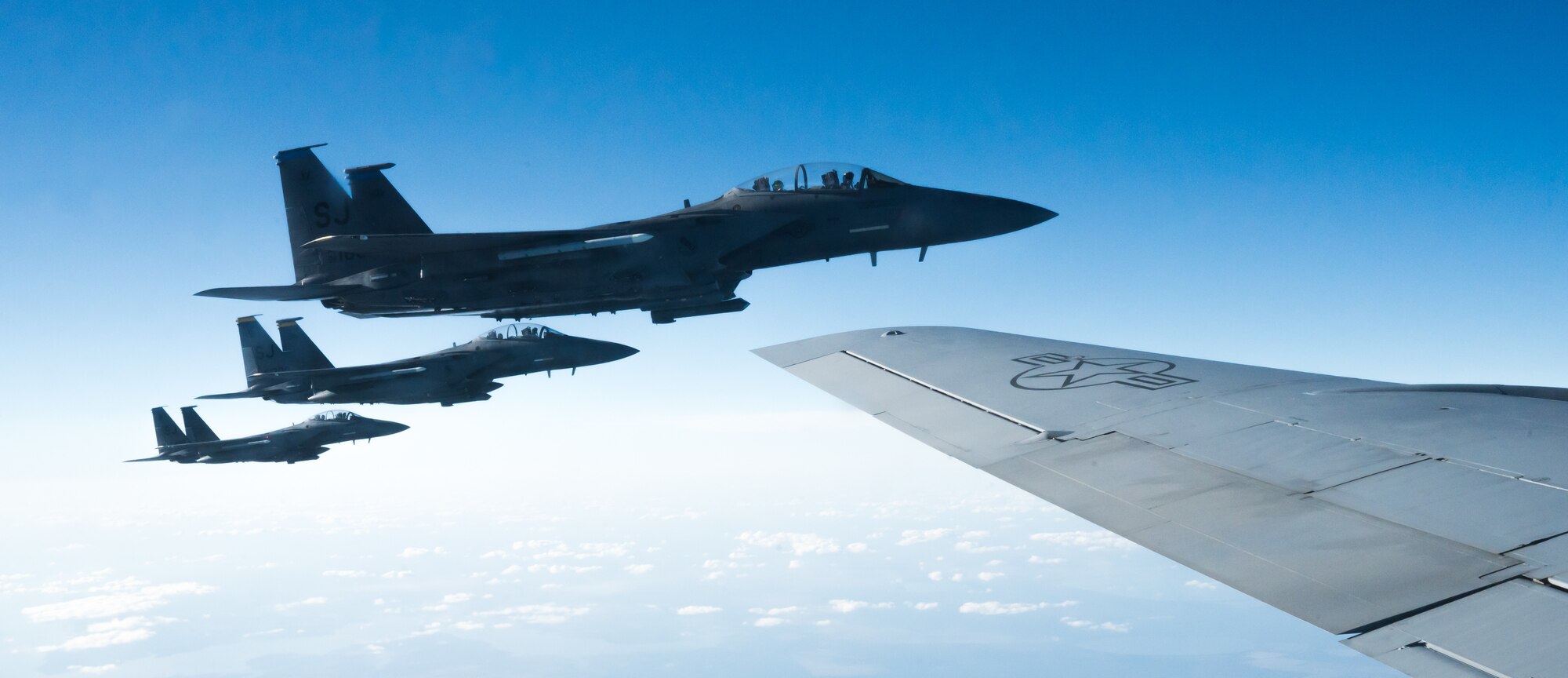 Three military jets fly in formation next to a fourth aircraft. The wing of the fourth is visible in the photo, at right. All four aircraft are flying over visible clouds.