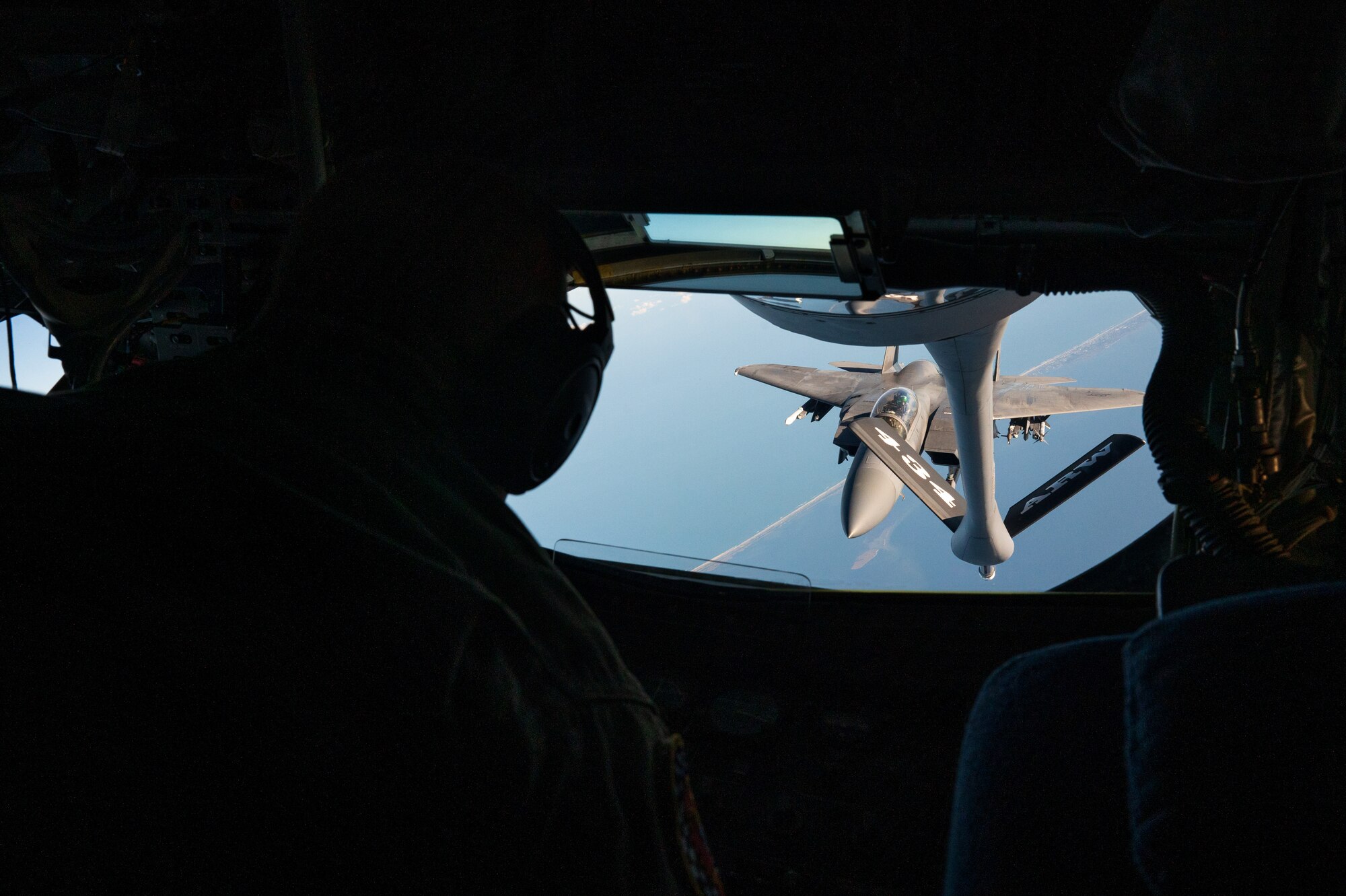 The silhouette of a man with a headset on looking out of a window. Out of the window, a military jet is visible as it flies over a coast.