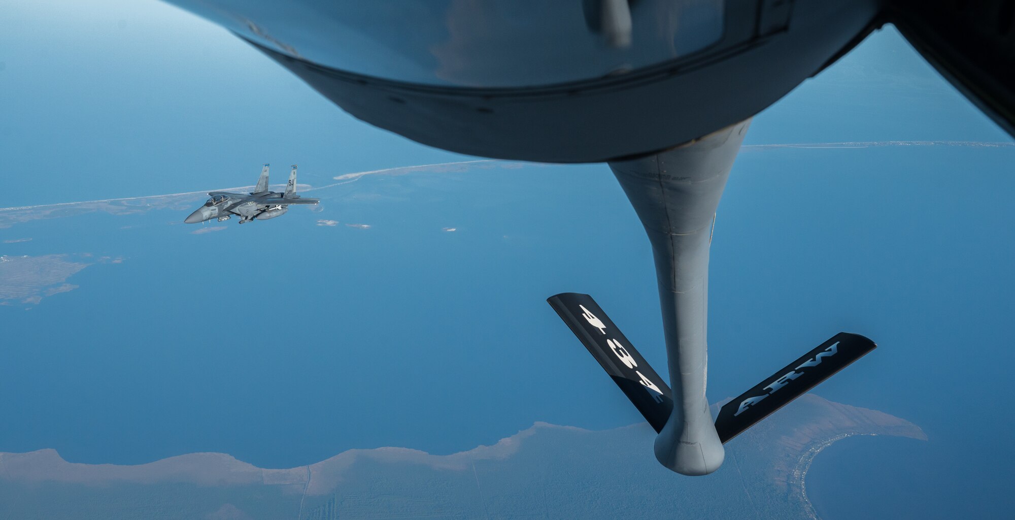 A military jet flies over a coast, at left of the frame. At right, the boom pod of another military aircraft is visible and it reads "434 ARW".
