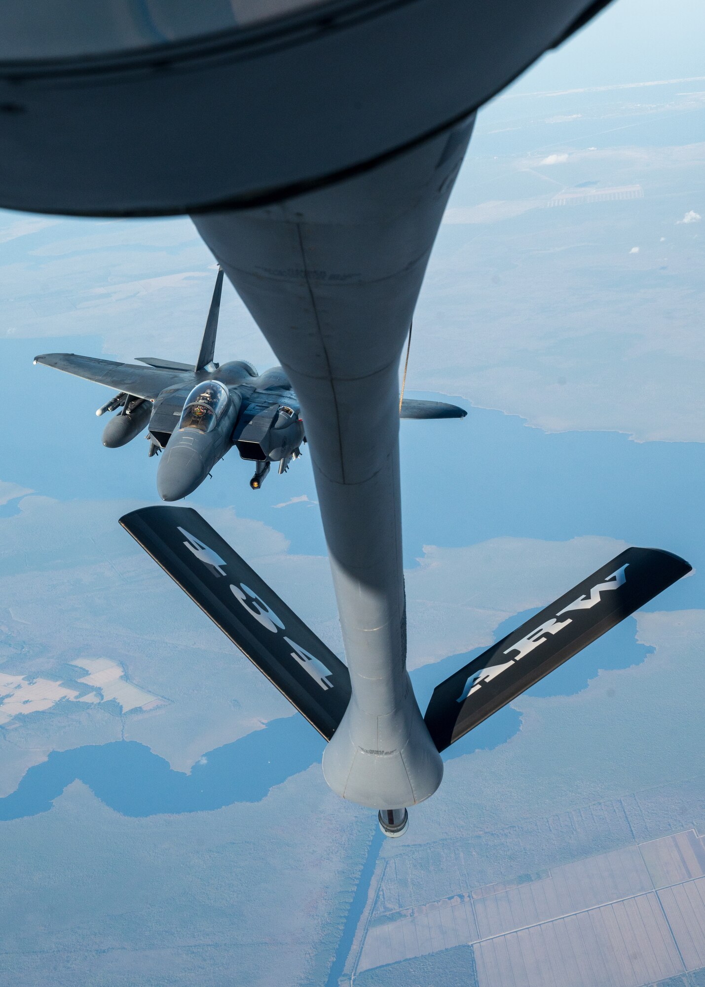 A military fighter jet flies over land and water. Another military aircraft's boom tail, visible in the foreground, reads "434 ARW".