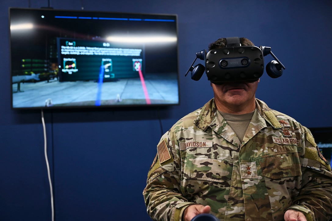 An airman uses virtual reality headset and joysticks in front of a TV screen with a virtual scenario.