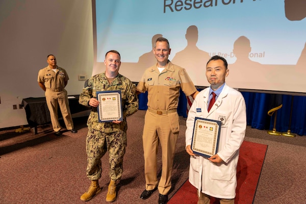 241010-JC800-1013 BETHESDA, Md. (Oct. 10, 2024) - Capt. William Deniston, lead inspector for the Medical Inspector General, awards a certificate of recognition to Cmdr. Noel Dickens and Dr. Jeffery Kim, program directors for research and integrity program,  during an outbrief for MEDIG. (U.S. Navy Photo by Mass Communication Specialist 1st Class Heath Zeigler)