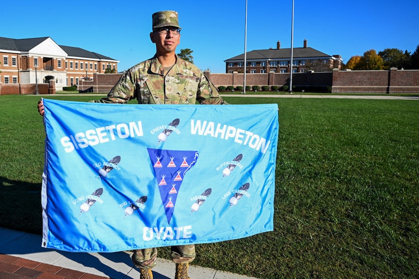 A service member holds a flag in front of a well-maintained lawn and buildings in the background.