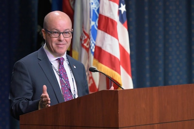 White man in a dark suit stands at a podium in front of flags