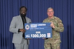 A black man in a grey suit and a white man in camo uniform hold a large sign of a check for $300K.
