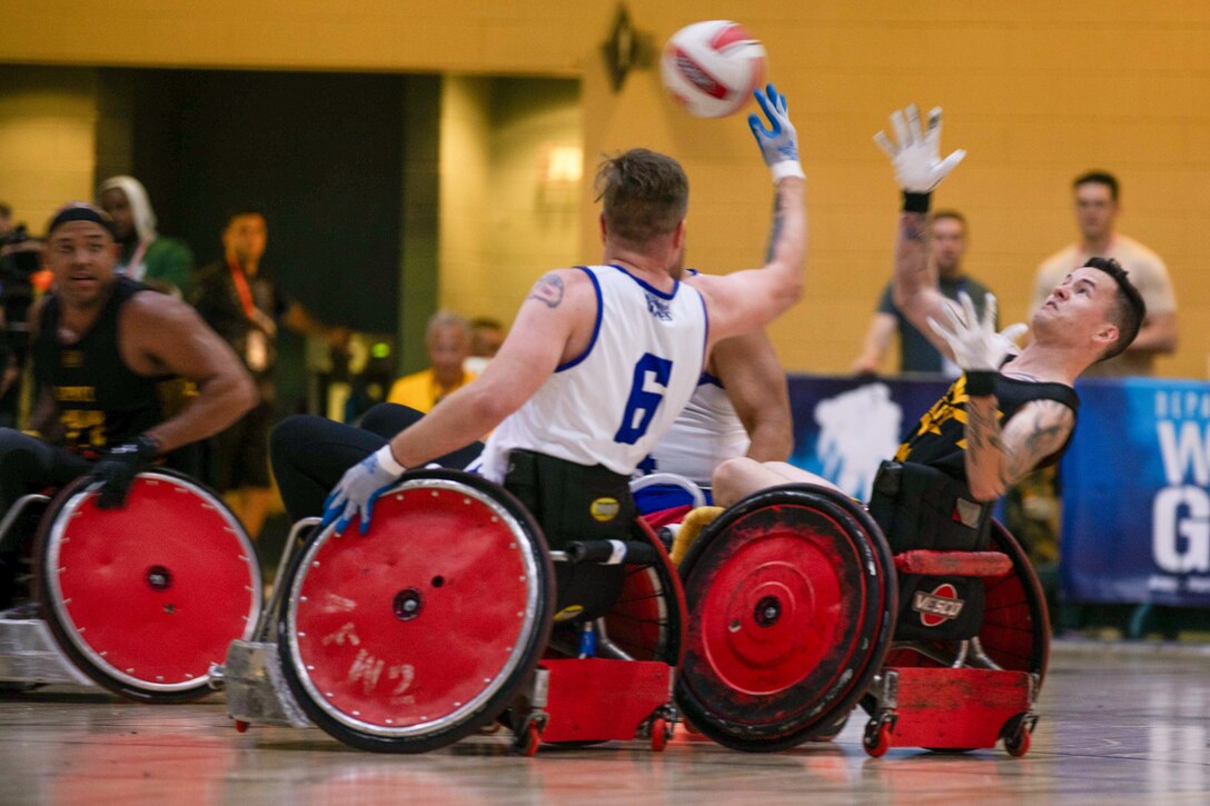 Two wheelchair rugby athletes - one in a black jersey; one in white  reach for a ball in play as a third looks on.