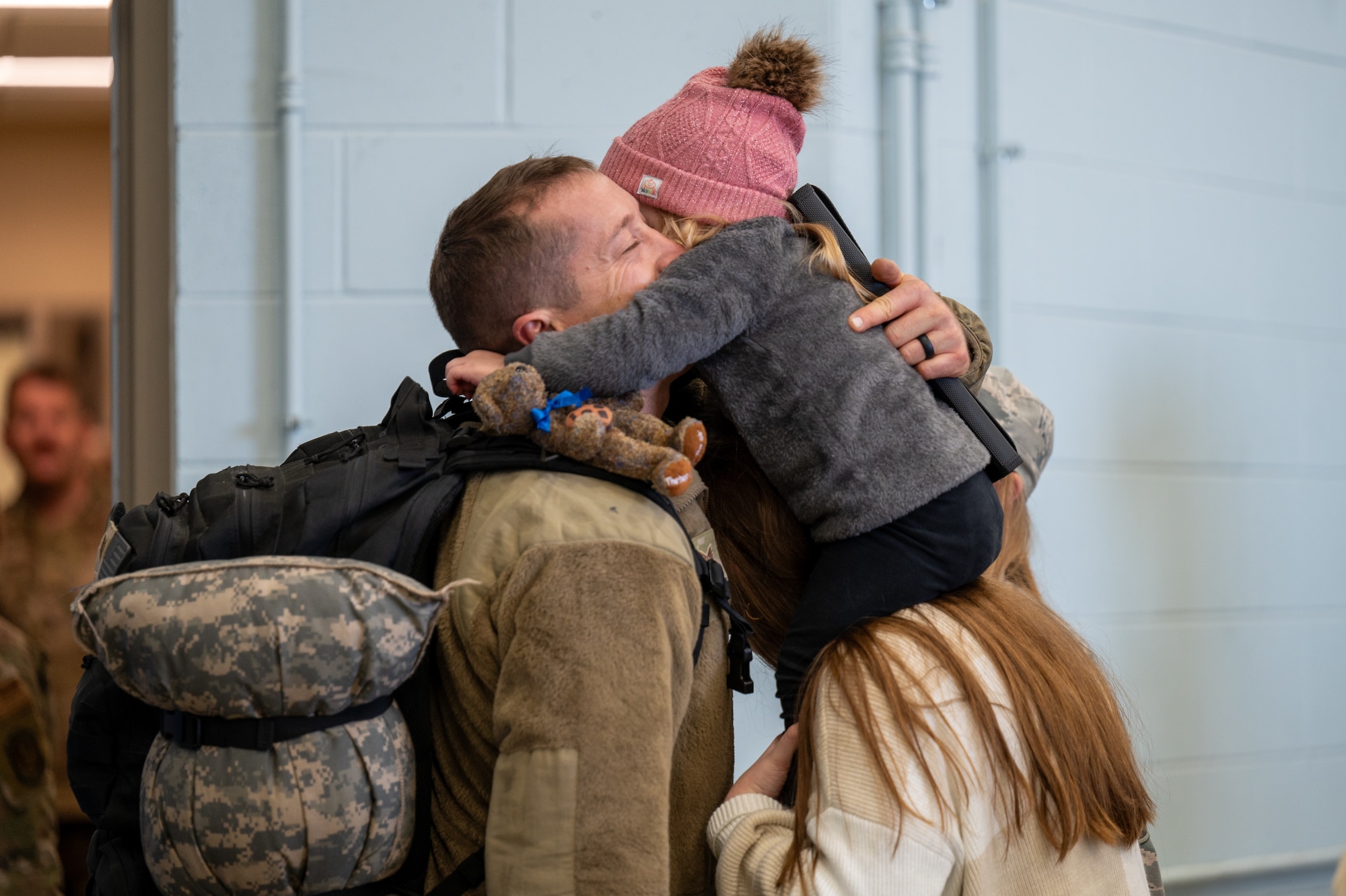 A man in a military uniform embraces his daughter. She is on the shoulders of a woman.
