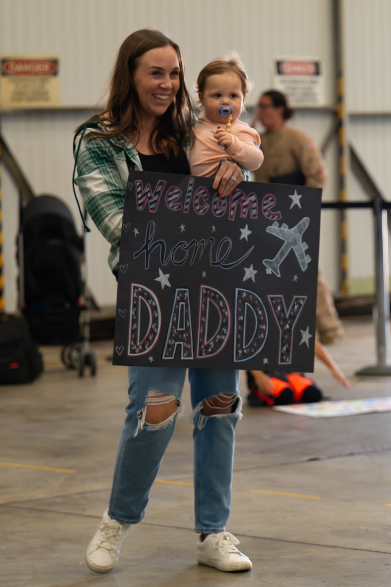 A woman holds her daughter as well as a sign that reads "Welcome home Daddy".