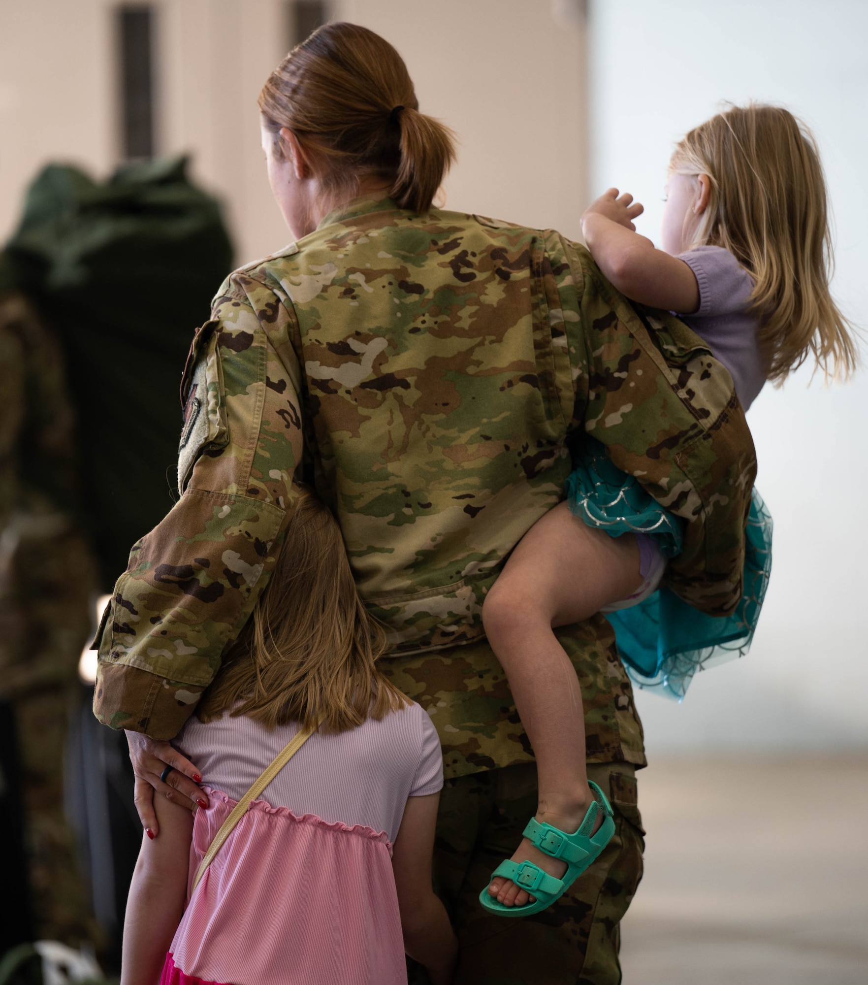 A woman in a military uniform holds her two children. One in her arms, the other at her side as they walk away from the camera.
