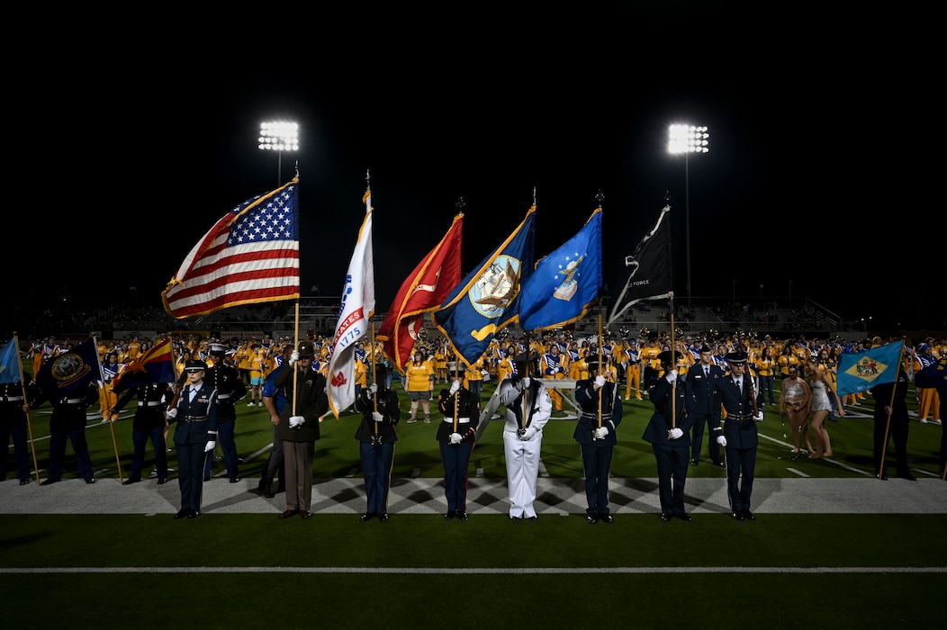 Service members hold flags of the armed forces and the U.S.A. flag at a football stadium.