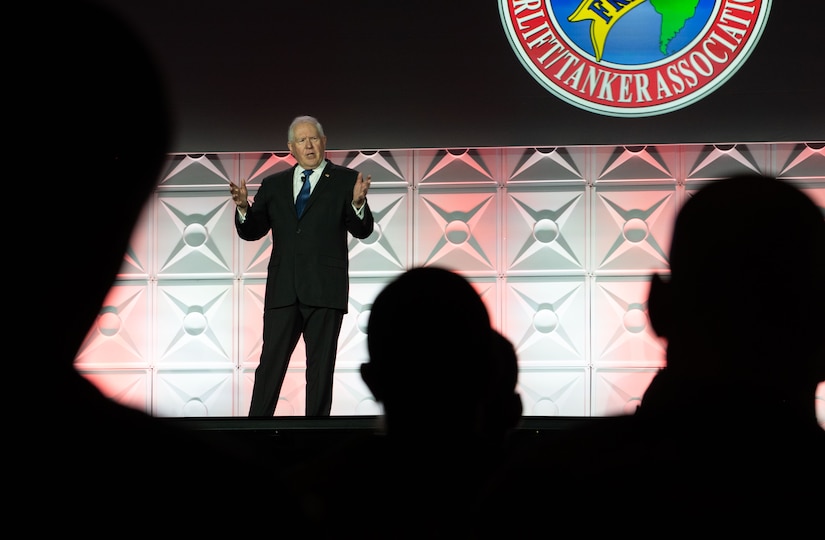 A man in a business suit stands in front of an audience on a stage.