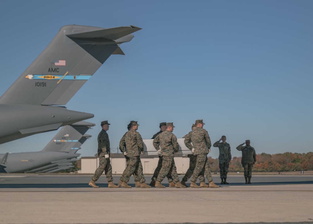 A group of Marines carry a transfer case on a flightline with aircraft in the background.