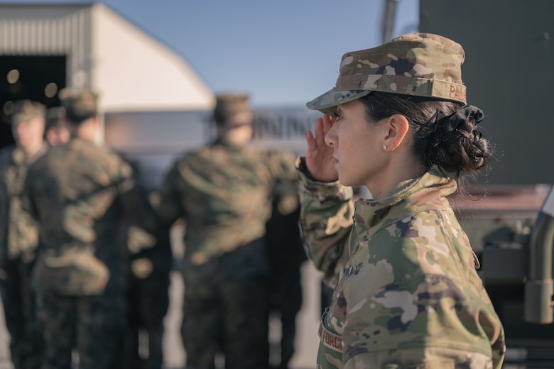 An Airman salutes as a group of Marines secure a transfer case in preparation to carry it to an awaiting transfer vehicle during a training event.