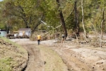 Two men walk down a dirt road with trees to their right, tree limbs on the ground and a house in the distance