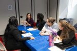 Five women sit at a table with blue table cloth talking.