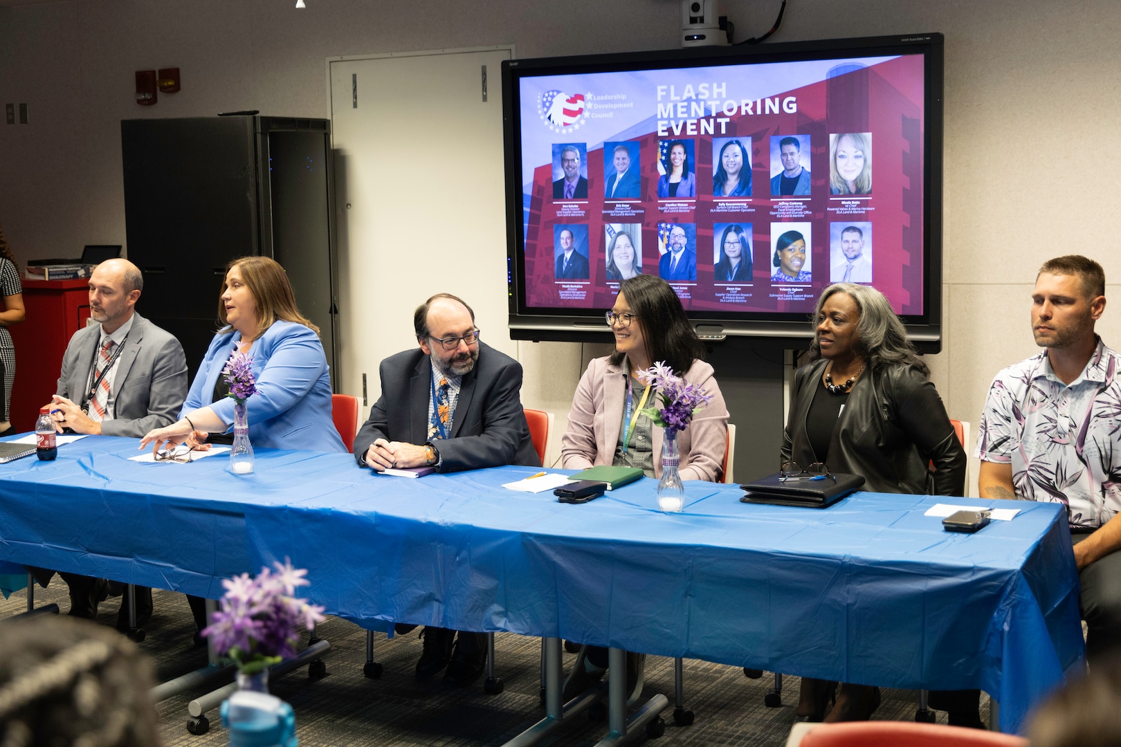 Six people seated at table with blue table cloth - 'Flash Mentoring Event' on TV in background.