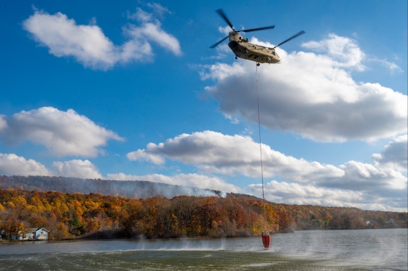 A helicopter carrying a bucket flies over a river.