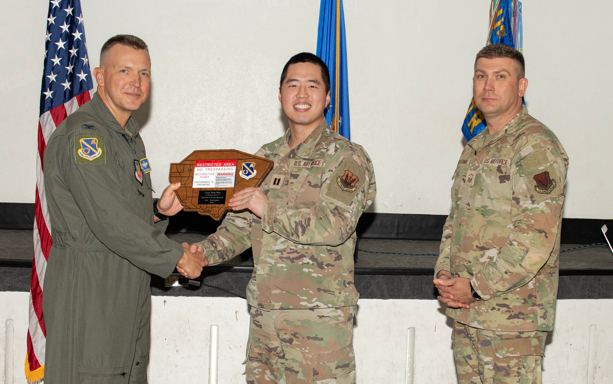 U.S. Air Force Col. Dane J. Gehrmann, left, commander of the Nevada Test and Training Range (NTTR), and Senior Master Sgt. Dennis Kolesnik, right, senior enlisted leader assigned to NTTR, present the Volunteer of the Quarter award to Capt. Yoon Kim, a project manager assigned to NTTR during a commander’s call at Nellis Air Force Base, Nevada, Oct. 31, 2024.