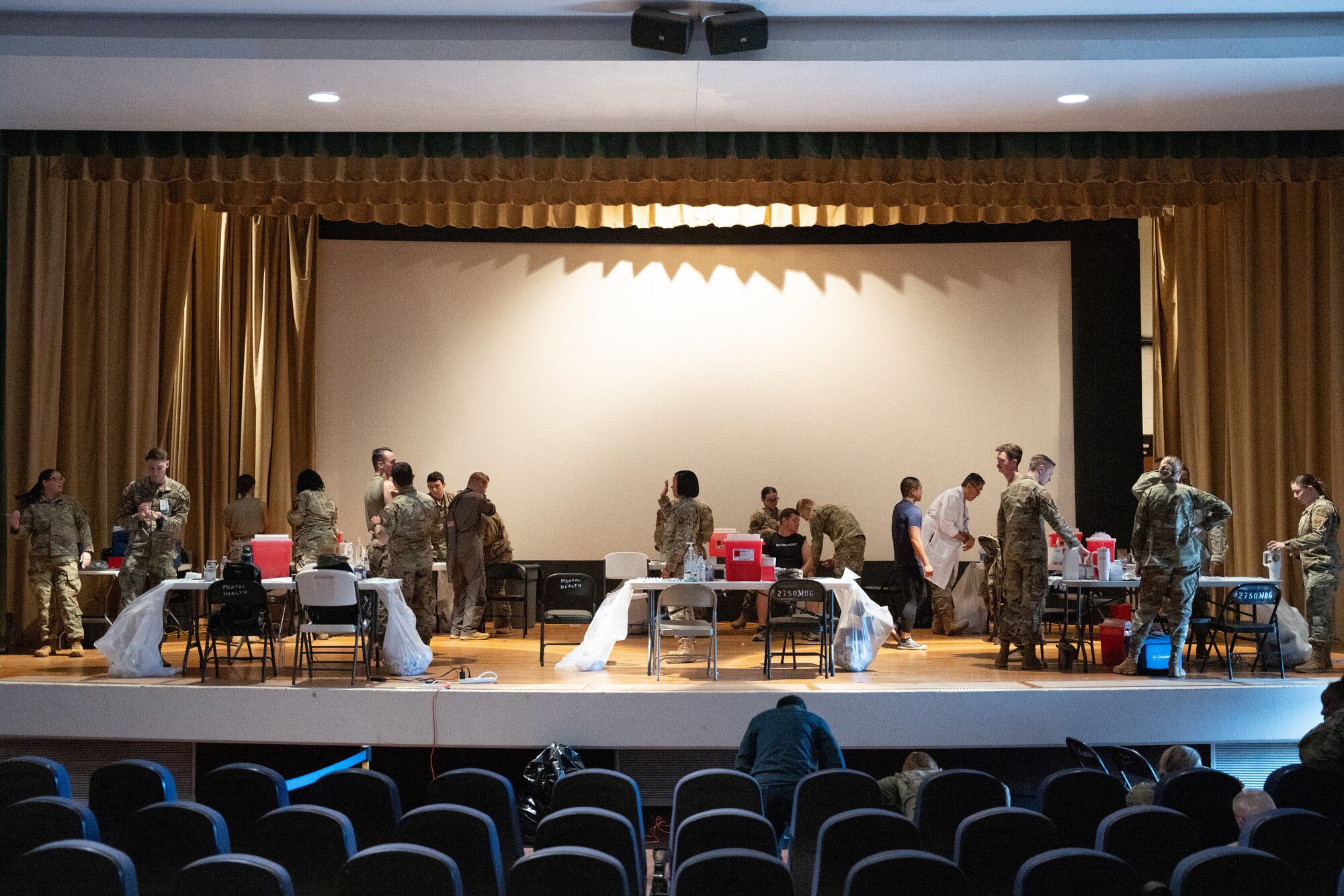 A group of individuals in military uniform and medical lab coats handle medical supplies on a stage filled with tables.