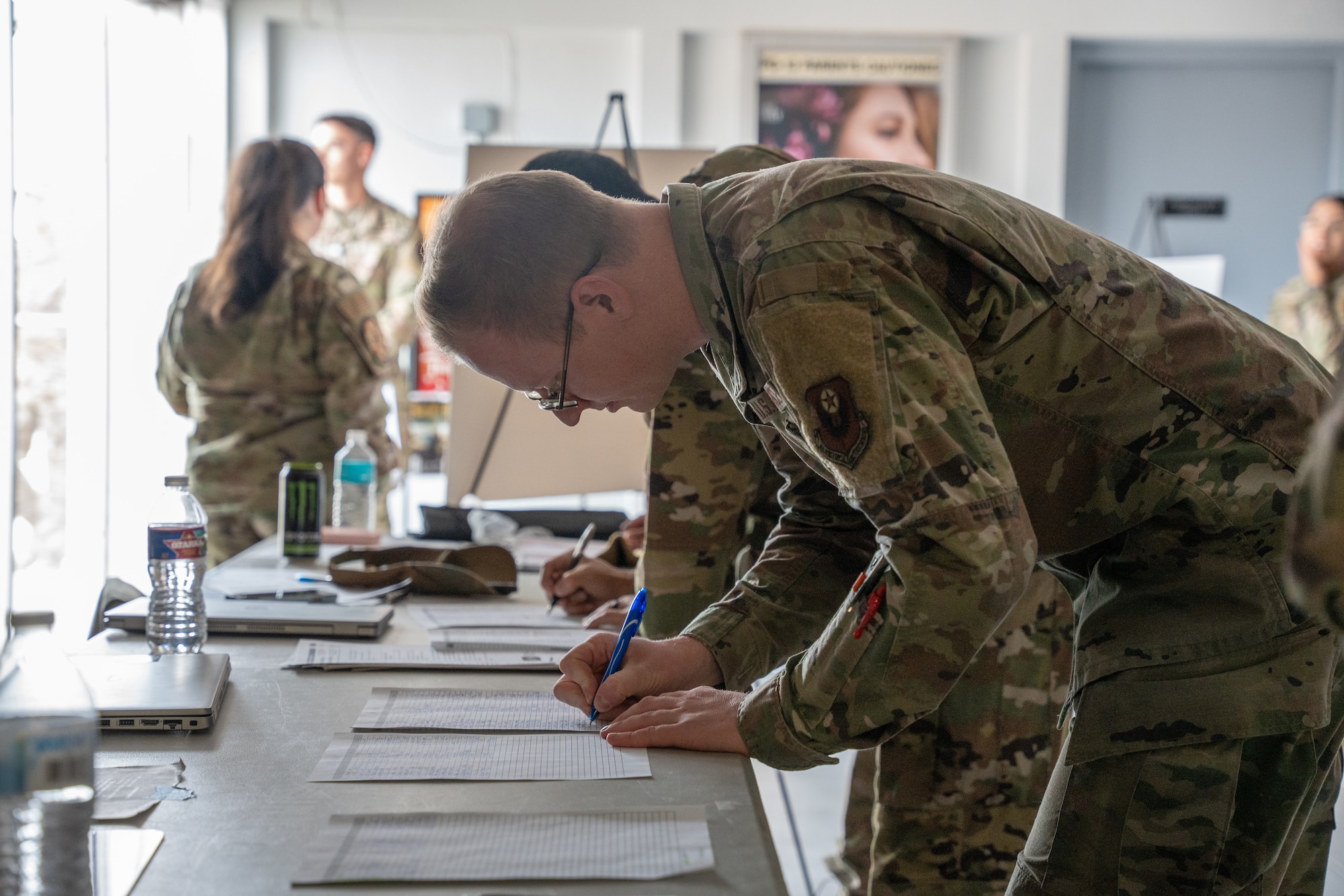 People in military uniform fill and sign rosters sitting on tables.