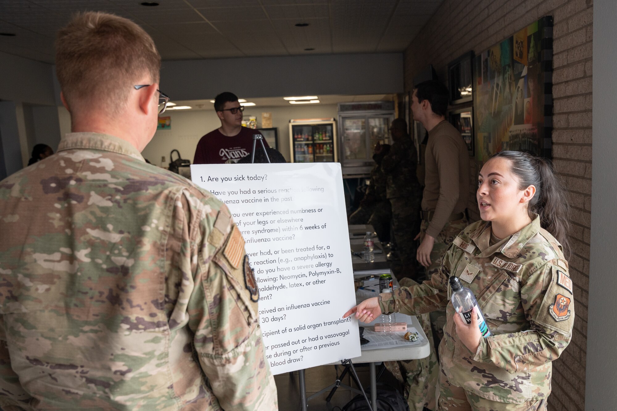 A women in military uniform speaks to a man in military uniform while she points at a poster with a list of medical questions.
