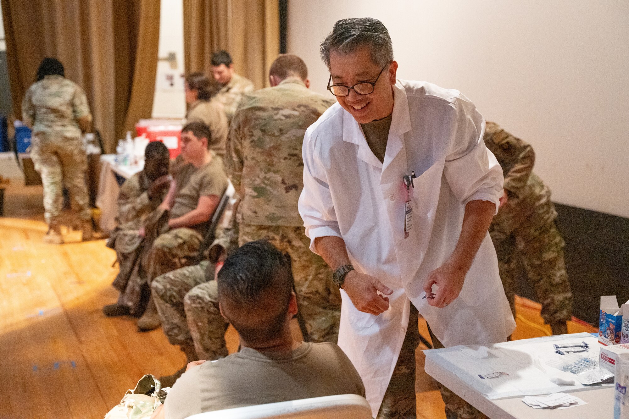 A man in a medical lab coat smiles at a seated person, while several people in military uniform work behind him.