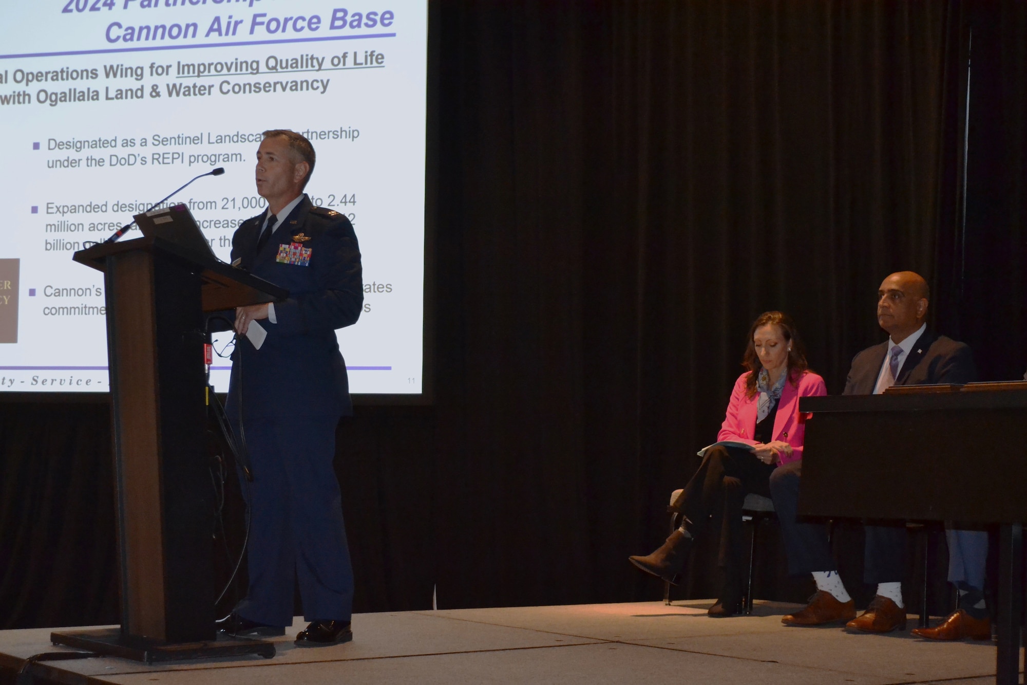 A man in military dress uniform speaks at a lectern with several individuals in business dress seated behind him.