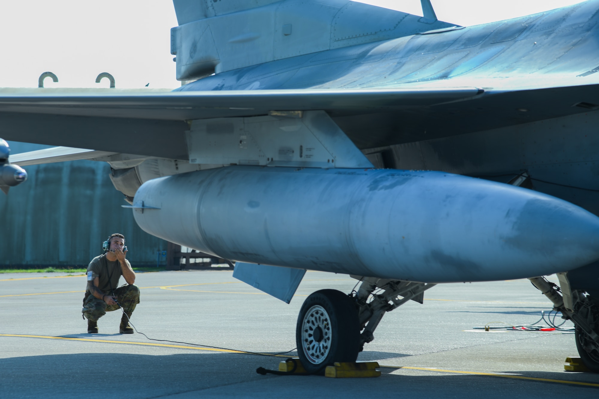 An Airman crouches behind an F-16C fighter jet and speaks into his headset microphone.