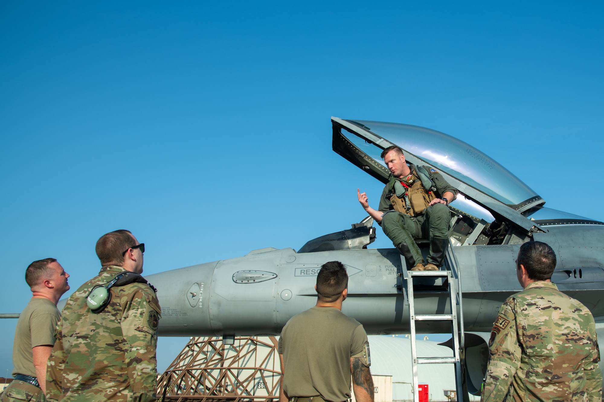 An F-16C fighter jet pilot sits on at the top of a ladder leading to the cockpit of an F-16C fighter jet. The pilot is looking down while speaking to four airmen as they look upwards and listen.
