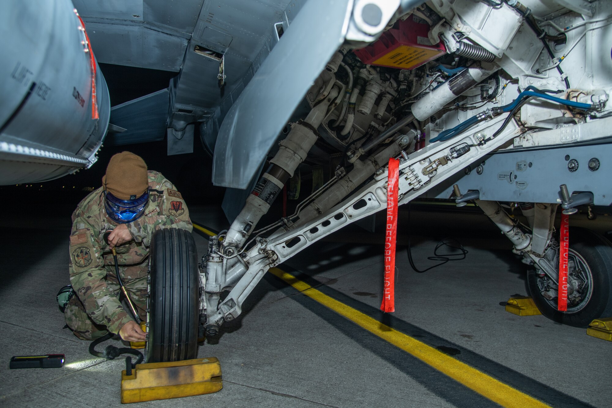 An Airman checks the tire pressure of an F-16C fighter jet, in the dark, illuminated by a flashlight.