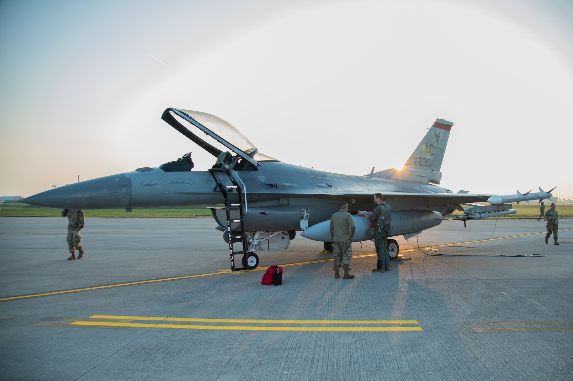 Two men stand in front of an F-16C fighter jet during the sunrise.