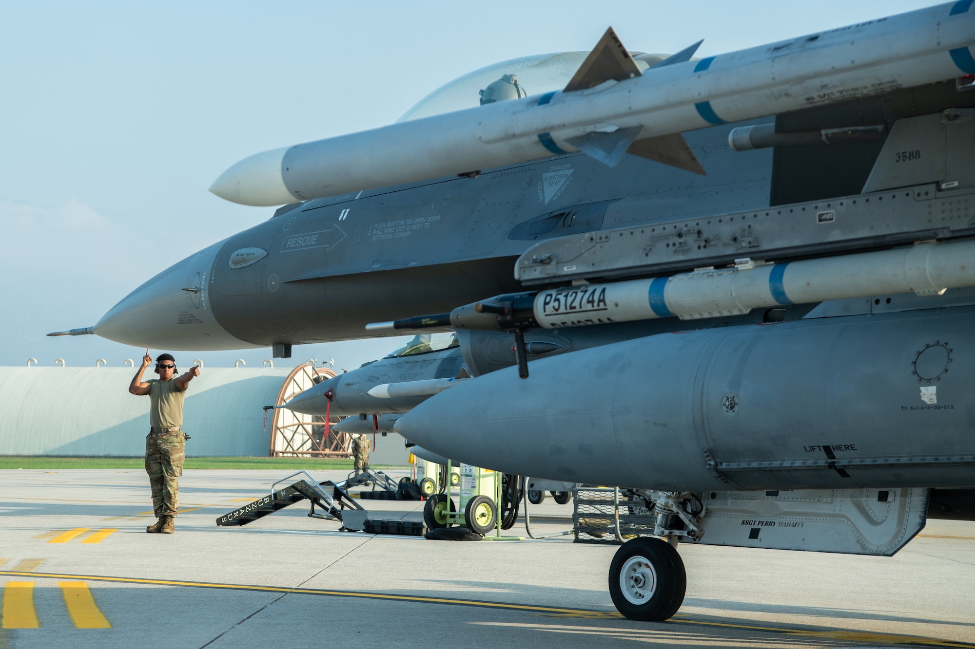 An Airman stands to the side of an F-16C fighter jet, pointing one hand at the jet.