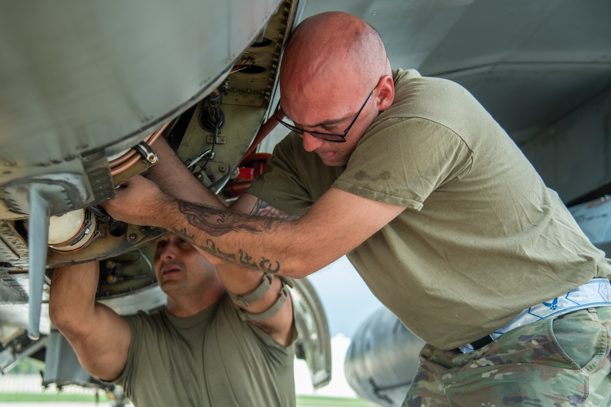 Two aircraft maintainers work underneath an F-16C fighter jet.